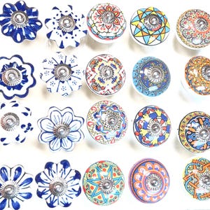 Op de afbeelding: Een verzameling van twintig decoratieve keramische kastknoppen. De knoppen hebben verschillende bloemen- en geometrische ontwerpen in blauw, wit, geel, oranje en rood. Elke knop heeft een zilverkleurige metalen basis en een uniek geschilderd patroon.