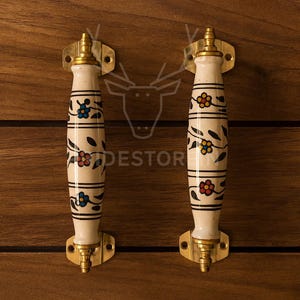 May include: Two decorative cabinet handles with a cream-colored, cylindrical design. Each handle features a floral pattern in blue, red, and yellow, with black accents. The handles are mounted on brass-colored hardware and set against a wooden background.