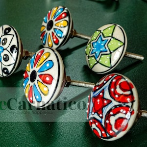 Może przedstawiać: Pięć ceramicznych gałek do szuflad z kolorowymi wzorami kwiatowymi i geometrycznymi. Gałki są białe z niebieskimi, czerwonymi, zielonymi, żółtymi i czarnymi akcentami. Każda gałka ma srebrną śrubę.