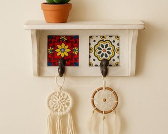 Puede incluir: Una estantería de madera blanca con dos baldosas de cerámica decorativas, cada una con un patrón floral diferente. Debajo, dos ganchos de metal oscuro, cada uno sosteniendo un atrapasueños. Una pequeña suculenta en maceta se encuentra en la estantería.