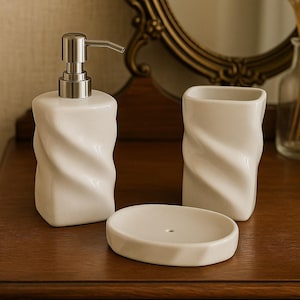 May include: A white ceramic bathroom set with a twisted design. The set includes a soap dispenser with a silver pump, a tumbler, and a soap dish. The items are arranged on a wooden surface.
