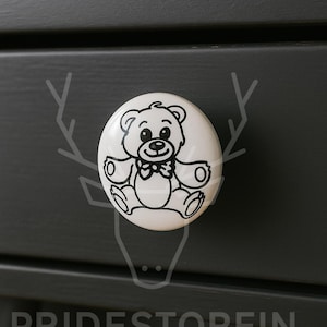 May include: A white drawer knob featuring a black outline of a teddy bear wearing a bow tie. The knob is attached to a dark grey drawer. The teddy bear design is in the centre of the knob.