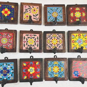 Könnte beinhalten: Set aus zwölf dekorativen Wandhaken. Jeder Haken hat eine quadratische, handbemalte Fliese mit einem Blumendesign in verschiedenen Farben, eingebettet in einen dunklen Holzrahmen. Die Haken sind schwarz und unten am Rahmen befestigt.