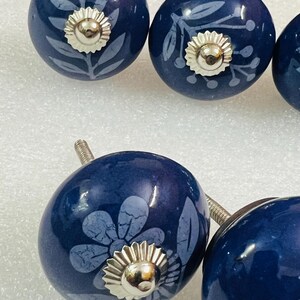 May include: A set of round, dark blue ceramic cabinet knobs featuring light blue botanical and floral patterns. Each knob is accented with a central, circular, silver-toned metal hardware piece and includes a threaded metal bolt for installation.