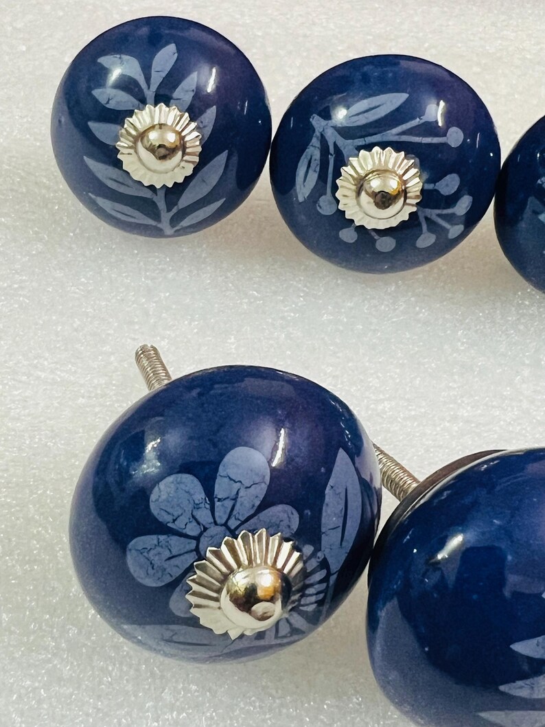 May include: A set of round, dark blue ceramic cabinet knobs featuring light blue botanical and floral patterns. Each knob is accented with a central, circular, silver-toned metal hardware piece and includes a threaded metal bolt for installation.