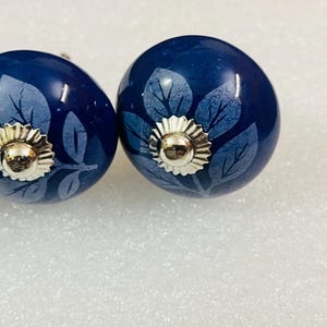 May include: Two round, dark blue ceramic cabinet knobs featuring a lighter blue leaf pattern. Each knob is centered with a polished, silver-toned metal cap that has a scalloped edge and a smooth, rounded dome in the middle.