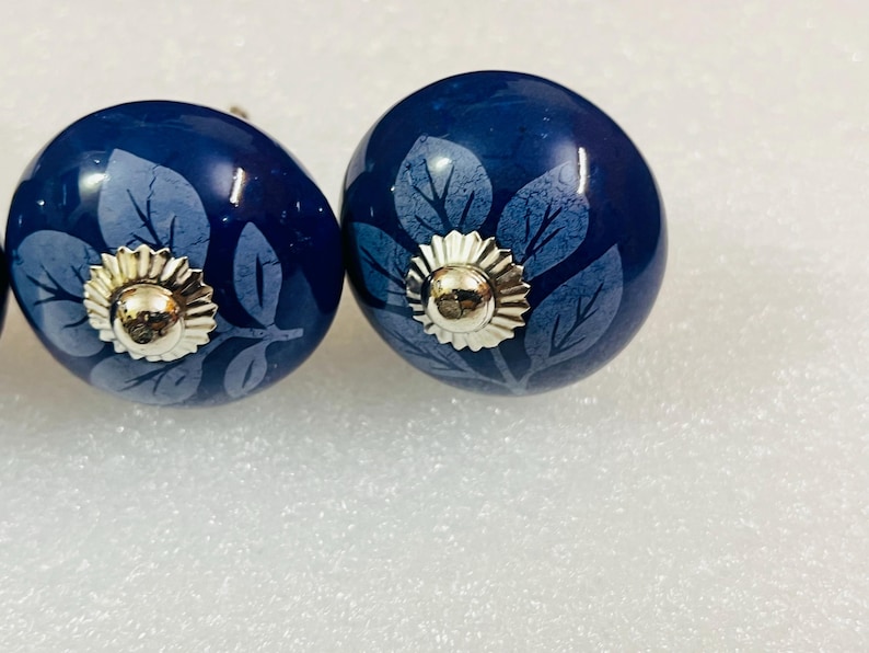 May include: Two round, dark blue ceramic cabinet knobs featuring a lighter blue leaf pattern. Each knob is centered with a polished, silver-toned metal cap that has a scalloped edge and a smooth, rounded dome in the middle.