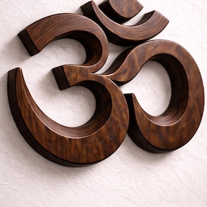 May include: A wooden Om symbol in a dark brown hue, showcasing the natural wood grain. The three-dimensional design is mounted on a textured white background. The symbol is a representation of a spiritual icon, with a contrasting white backdrop.