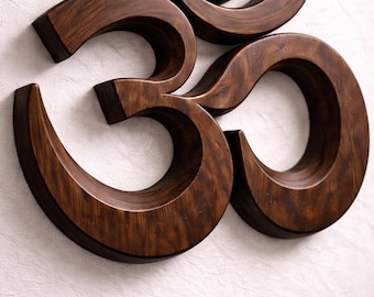 May include: A three-dimensional Om symbol crafted from dark brown wood with a visible grain pattern. The wall-mounted piece features smooth, curved edges and a polished finish, showcasing the iconic sacred character in a bold, raised design.