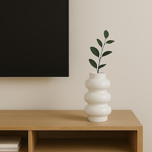 May include: A white, bulbous ceramic vase with a dark green leafy stem. The vase has a unique, stacked design and sits on a light brown wooden surface. The background is a neutral beige.
