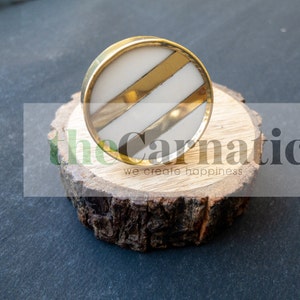 May include: A round, white and gold cabinet knob with two gold stripes. The knob is sitting on a piece of wood.