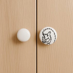 May include: Two white cabinet knobs. One is a simple, round design. The other is a white knob with a black and white illustration of a hedgehog. The knobs are mounted on a light-coloured wooden cabinet door.