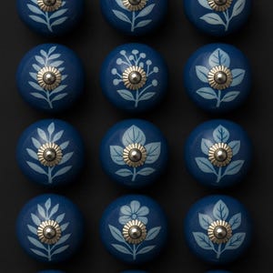 Peut inclure: Douze boutons de meuble bleu marine. Chaque bouton présente un motif floral blanc et un centre en métal doré. Les boutons sont disposés en grille sur un fond sombre, mettant en valeur leur aspect décoratif et leur design floral.