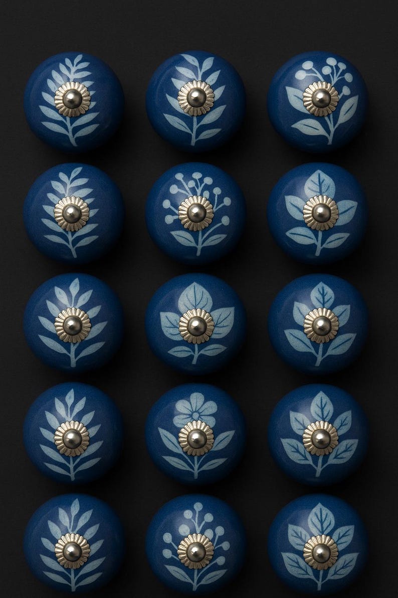 May include: A set of fifteen round, dark blue ceramic cabinet knobs arranged in a grid. Each knob features a light blue botanical print of leaves, flowers, or berries and a central silver-toned metal screw head with a ridged edge.