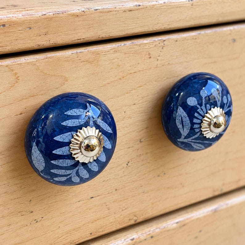 May include: Two round, dark blue ceramic cabinet knobs featuring a light blue leaf and floral pattern. Each knob is centered with a shiny, metallic gold-toned circular plate and a small, raised gold-toned center stud for attachment.