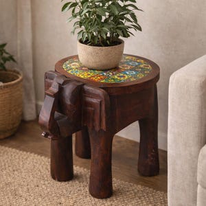 May include: A dark brown, hand-carved wooden elephant stool with a round top featuring a colorful mosaic tile design. A potted plant sits on top of the stool. The stool has four legs and is placed on a woven rug.