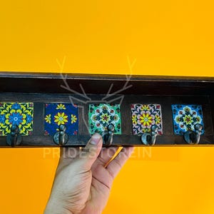 Może przedstawiać: Drewniany wieszak ścienny z pięcioma dekoracyjnymi płytkami ceramicznymi. Każda płytka ma unikalny wzór kwiatowy w żywych kolorach. Wieszak ma pięć metalowych haczyków. Widoczny jest tekst "PRIDESTOREIN".