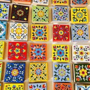 Op de afbeelding: Een verzameling kleurrijke, vierkante keramische tegels met bloempatronen. De tegels hebben verschillende ontwerpen in tinten blauw, geel, rood, oranje en groen. Elke tegel heeft een uniek bloemmotief, wat een levendige en decoratieve weergave creëert.