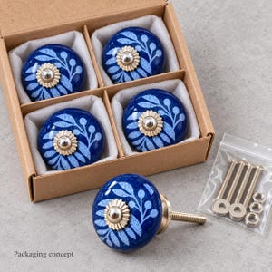May include: A set of five round ceramic cabinet knobs featuring a deep blue base with a light blue leaf and berry pattern. Each knob has a central silver-toned metal accent. Four knobs are nestled in a cardboard box, with one placed outside.