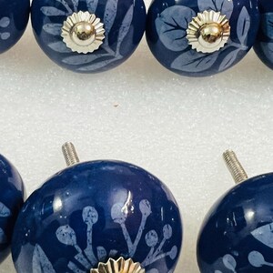 May include: A set of round, dark blue ceramic cabinet knobs featuring light blue botanical patterns of leaves and berries. Each knob has a polished silver-tone metal center cap and a threaded metal bolt extending from the back for installation.