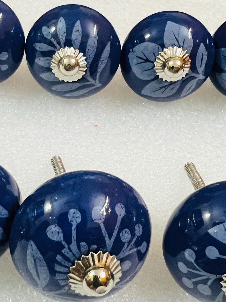 May include: A set of round, dark blue ceramic cabinet knobs featuring light blue botanical patterns of leaves and berries. Each knob has a polished silver-tone metal center cap and a threaded metal bolt extending from the back for installation.