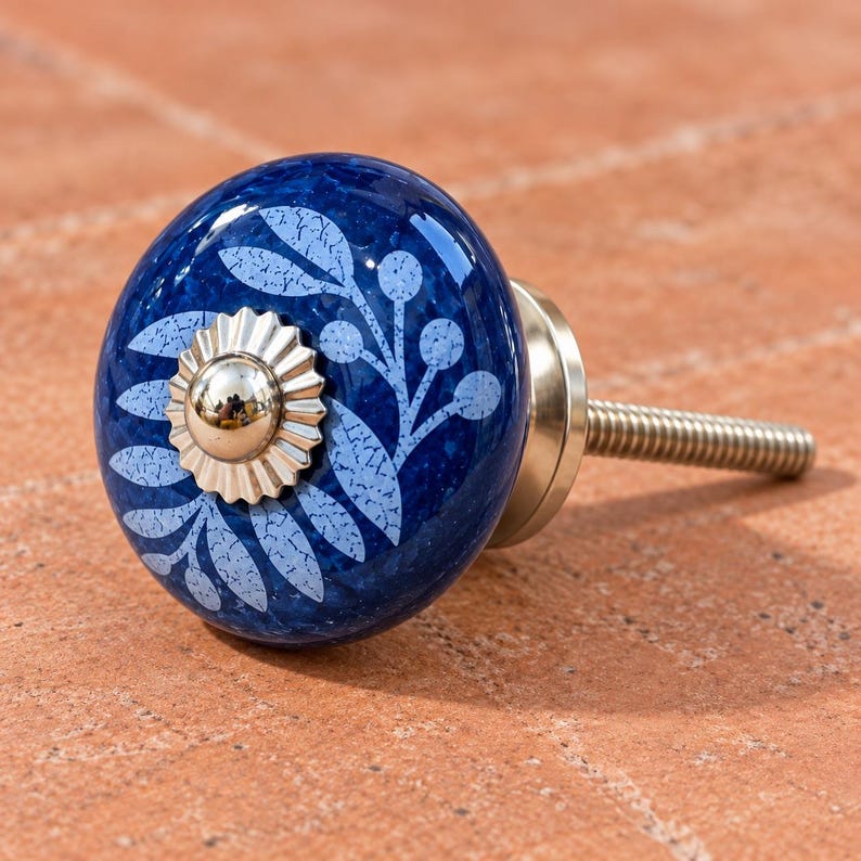 May include: A round, deep blue ceramic cabinet knob featuring a light blue botanical leaf and berry pattern. The center of the knob is adorned with a shiny, scalloped silver-tone metal cap, and it includes a threaded metal bolt for installation.