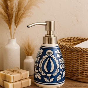Könnte beinhalten: Blau-weißer Keramik-Seifenspender mit floralem Muster und silberner Pumpe. Auf dem Bild sind auch verpackte Seifenstücke, dekorative Vasen und ein Weidenkorb zu sehen. Ein stilvolles Badezimmer-Accessoire.
