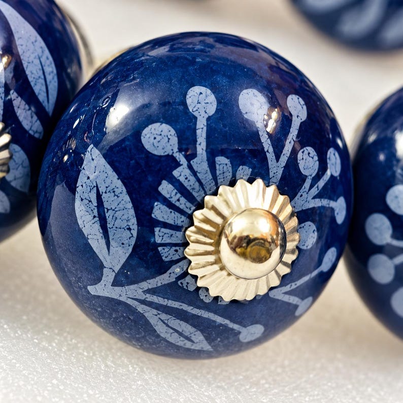 May include: A round, dark blue ceramic cabinet knob featuring a light blue, textured floral and leaf pattern. The center of the knob is adorned with a shiny, scalloped gold-tone metal cap that secures the piece in place.