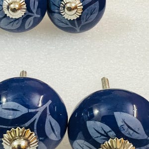 May include: Four round, dark blue ceramic cabinet knobs featuring a light blue leaf pattern. Each knob has a polished silver-tone metal center cap with a scalloped edge and a threaded metal bolt extending from the back for installation.