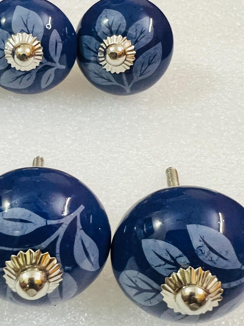 May include: Four round, dark blue ceramic cabinet knobs featuring a light blue leaf pattern. Each knob has a polished silver-tone metal center cap with a scalloped edge and a threaded metal bolt extending from the back for installation.