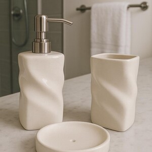 May include: A cream-colored bathroom set featuring a soap dispenser with a silver pump, a tumbler, and a soap dish. The set has a textured, twisted design. The items are arranged on a marble surface.