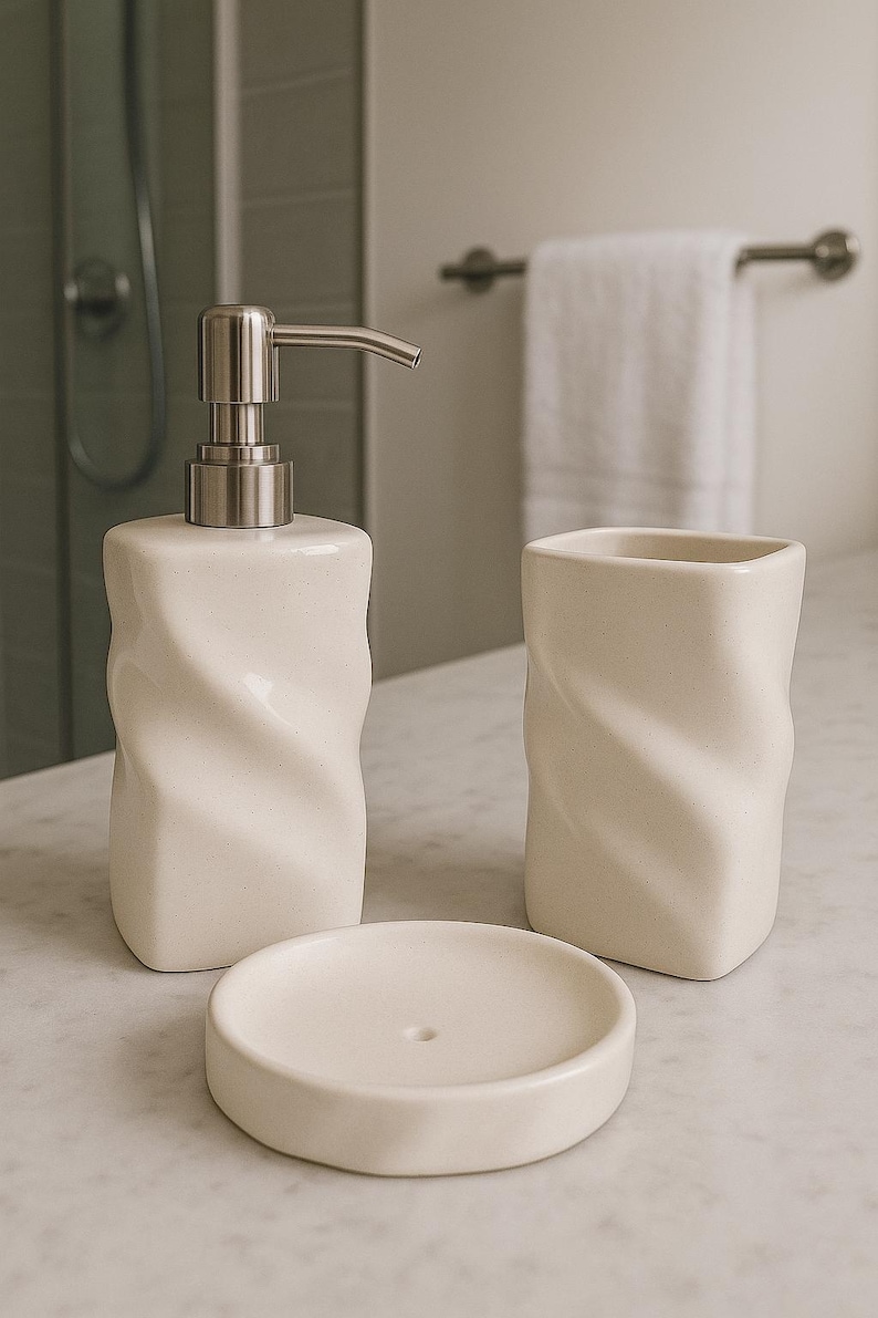 May include: A cream-colored bathroom set featuring a soap dispenser with a silver pump, a tumbler, and a soap dish. The set has a textured, twisted design. The items are arranged on a marble surface.