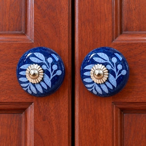 May include: Two round cabinet knobs featuring a deep blue glazed finish with a light blue leaf and berry pattern. Each knob has a central metallic gold-toned fluted circular accent piece mounted on a wooden cabinet door surface.