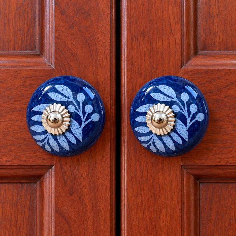 May include: Two round cabinet knobs featuring a deep blue glazed finish with a light blue leaf and berry pattern. Each knob has a central metallic gold-toned fluted circular accent piece mounted on a wooden cabinet door surface.