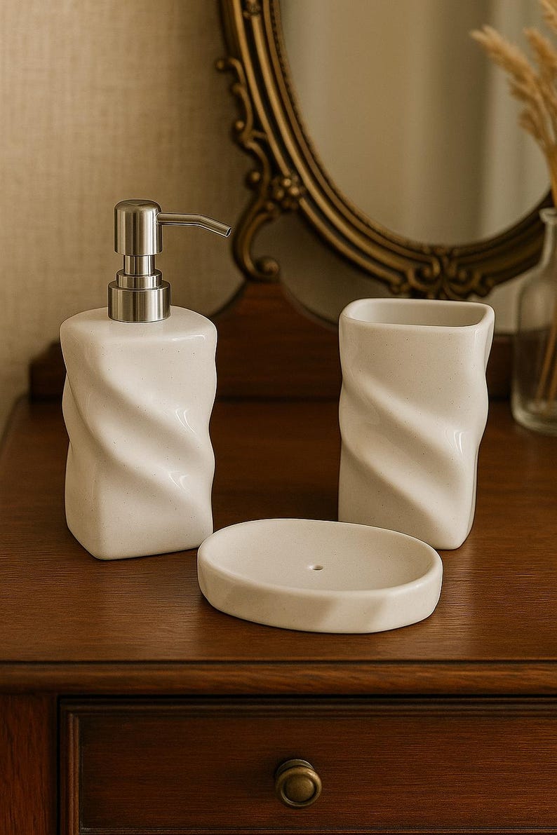 May include: A white ceramic bathroom set with a twisted design. The set includes a soap dispenser with a silver pump, a tumbler, and a soap dish. The items are arranged on a wooden surface.