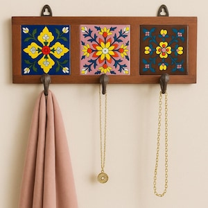 Puede incluir: Un perchero de pared de madera con tres baldosas de cerámica decorativas con diseños florales en amarillo, azul y rosa. El perchero tiene tres ganchos de metal y sostiene una bufanda rosa y dos collares dorados.