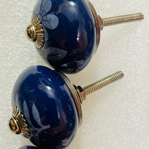 May include: Three round, dark blue ceramic cabinet knobs featuring a subtle, light-colored floral pattern. Each knob has a polished gold-tone metal base and a threaded metal bolt extending from the back for installation purposes.