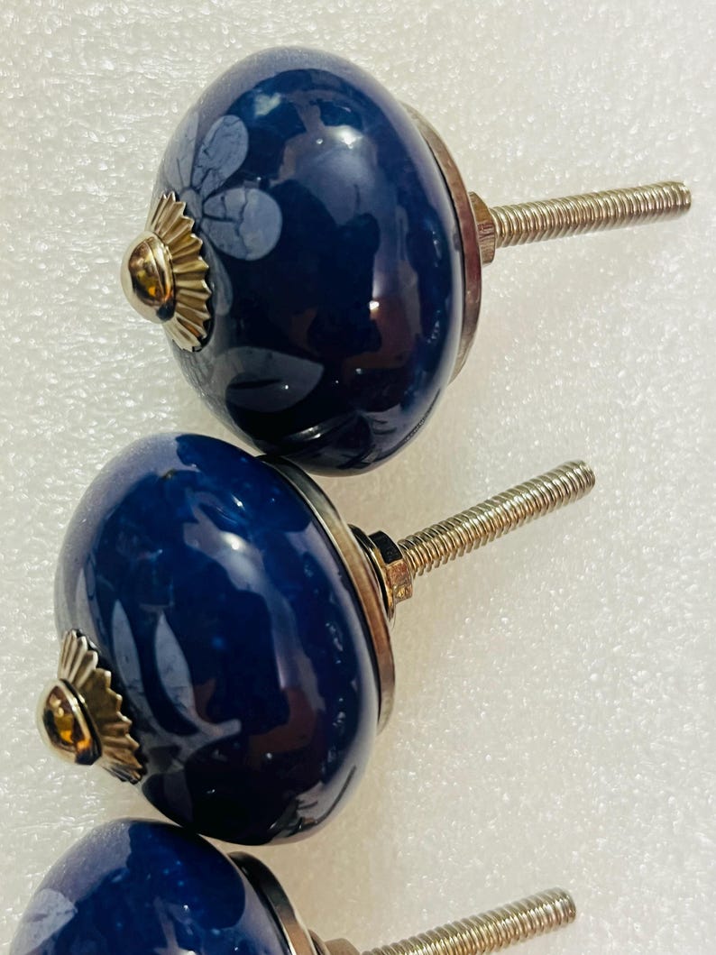 May include: Three round, dark blue ceramic cabinet knobs featuring a subtle, light-colored floral pattern. Each knob has a polished gold-tone metal base and a threaded metal bolt extending from the back for installation purposes.