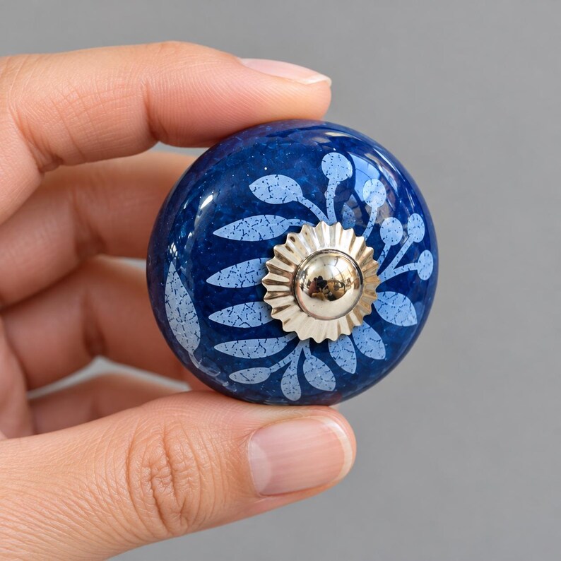 May include: A round, dark blue ceramic cabinet knob featuring a light blue, speckled floral pattern. The center of the knob is adorned with a shiny, silver-toned metal rosette with a scalloped edge and a smooth, reflective dome.