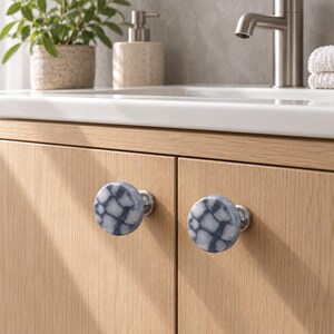May include: Two round cabinet knobs featuring a blue and white mosaic-style pattern with a cracked stone appearance. Each knob is mounted on a circular silver-toned metal base and attached to light-coloured wooden cabinet doors.