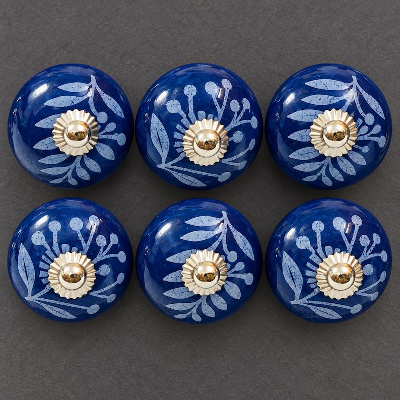 May include: A set of six round, dark blue ceramic cabinet knobs featuring a light blue botanical leaf and berry pattern. Each knob is centered with a polished, scalloped silver-tone metal hardware piece with a smooth, rounded top.