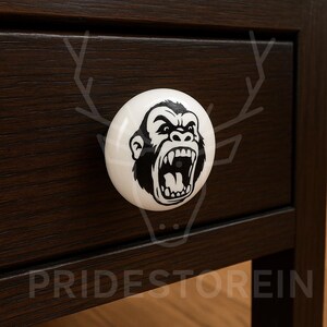 Pode incluir: Puxador de gaveta redondo de cerâmica branca com um desenho de rosto de gorila preto. O puxador está montado numa gaveta de madeira castanha escura. O texto "PRIDESTOREIN" é visível na parte inferior da gaveta.
