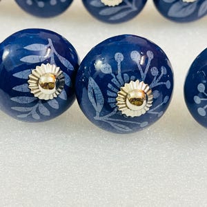 May include: A set of round, dark blue ceramic cabinet knobs featuring a light blue botanical print of leaves and berries. Each knob is finished with a polished silver-tone metal base and a central silver-tone decorative cap.
