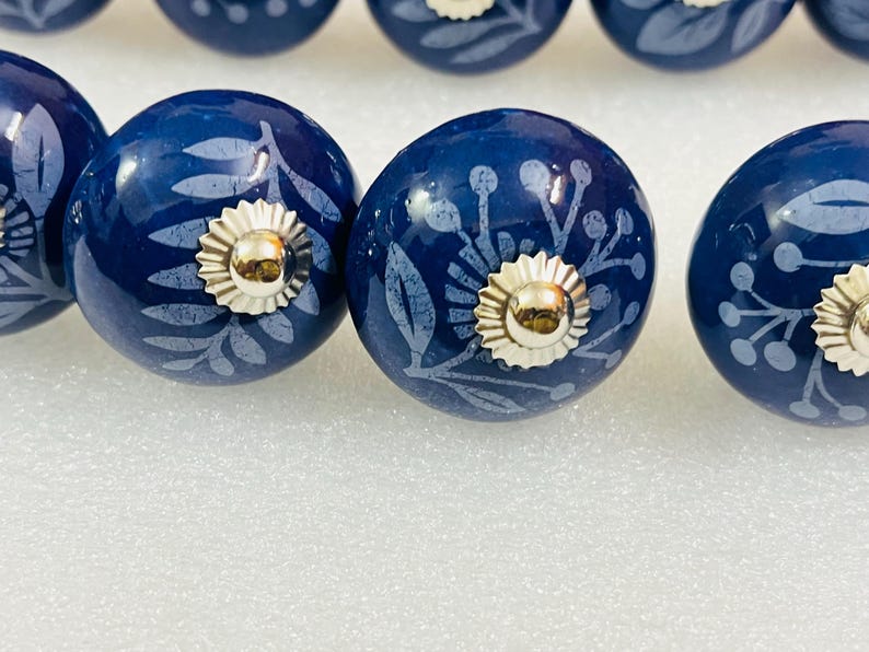 May include: A set of round, dark blue ceramic cabinet knobs featuring a light blue botanical print of leaves and berries. Each knob is finished with a polished silver-tone metal base and a central silver-tone decorative cap.