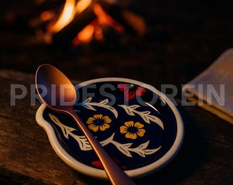May include: A copper-colored spoon rests on a ceramic spoon rest with a dark blue floral design. The spoon rest has a white rim and a curved handle rest. The background includes a blurred campfire and the text "PRIDESTOREIN".