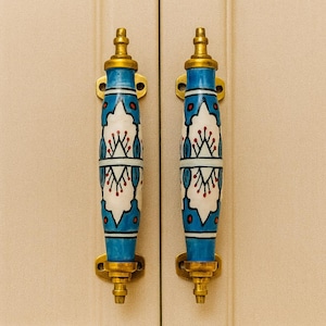 Peut inclure: Deux poignées de porte d'armoire avec un design bleu, blanc et doré. Les poignées sont cylindriques avec une base bleue, un centre blanc avec un motif floral et des accents dorés. Elles sont montées sur une porte d'armoire beige clair.
