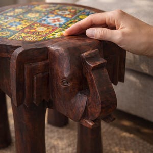 May include: A dark brown wooden stool carved in the shape of an elephant with a round top surface featuring a grid of colourful square tiles with floral patterns in shades of yellow, blue, red, and white, supported by four sturdy cylindrical legs.