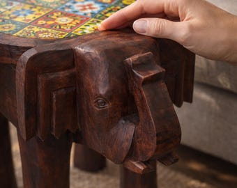 May include: A dark brown wooden stool carved in the shape of an elephant with a round top surface featuring a grid of colorful square tiles with floral patterns in shades of yellow, blue, red, and white, supported by four sturdy cylindrical legs.