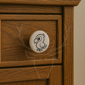 May include: A white cabinet knob with a black cartoon parrot design. The knob is round and attached to a wooden cabinet with a natural wood grain finish. The parrot has a large eye and a curved beak.