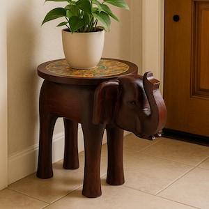 Puede incluir: Un soporte para plantas de madera marrón oscuro con forma de elefante y una parte superior redonda y estampada. Una maceta blanca con una planta verde encima. El soporte tiene cuatro patas y está colocado sobre un suelo embaldosado.